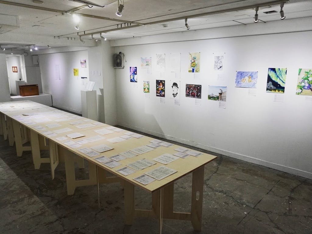 第1回刑務所アート展の展示会場。テーブルにエッセイや詩などの投稿作品、壁面に絵画作品を飾っている